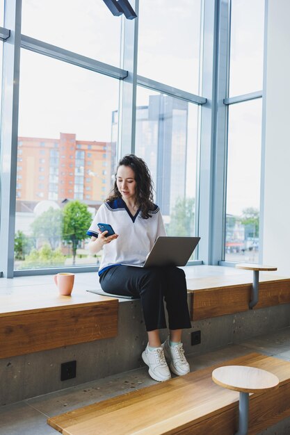 Een lachende jonge vrouw met krullend haar zit bij de grote ramen in een café en werkt op een laptop Werk op afstand