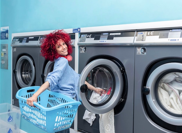 Een lachende jonge vrouw met rood afrohaar die de was doet in een blauwe automatische wasserette