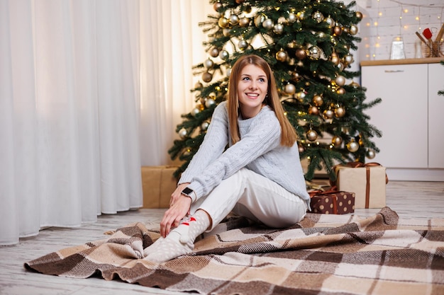 Een lachende jonge vrouw zit in de buurt van een kerstboom te versieren Gezellige kerstsfeer in huis