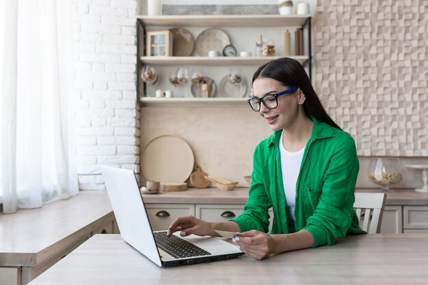 Een lachende jonge vrouw zit thuis in de keuken op een laptop te werken met een creditcard