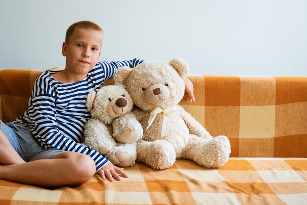 Een lachende jongen in gestreept t-shirt en blauwe korte broek zit op de bank en knuffelt knuffelbeer schattig portret van...