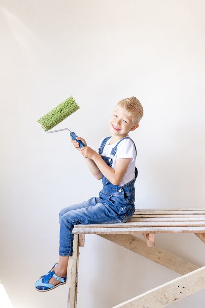 Een lachende kind bouwer zit op een bouwladder in een appartement met witte muren en houdt een kap voor het schilderen van muren, een plek voor tekst, een reparatie-concept