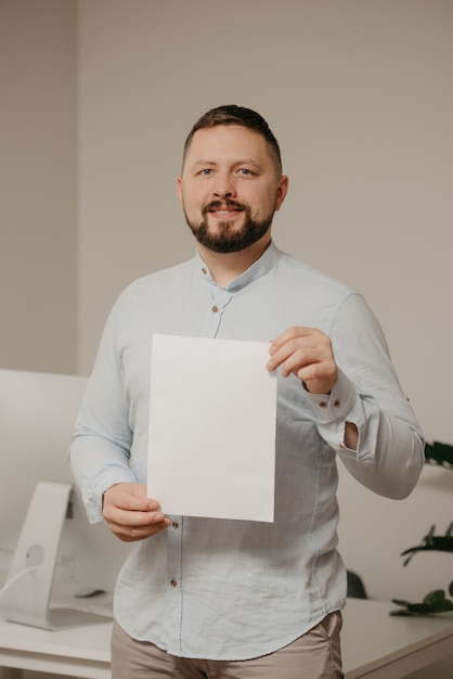 Een lachende man met een baard staat thuis met een leeg vel wit papier bij een stalen desktopcomputer