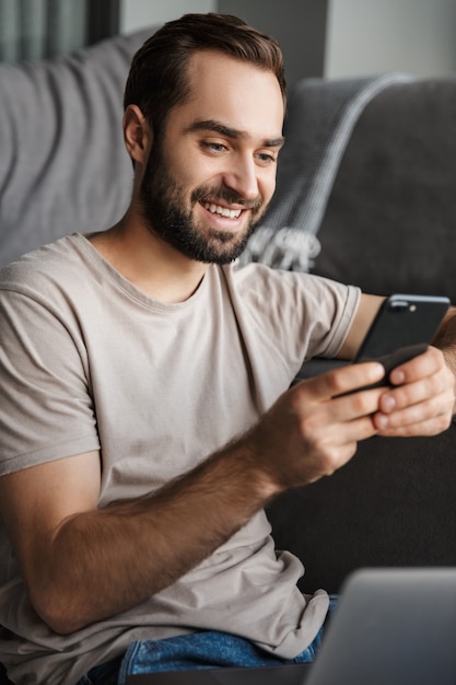 een lachende positieve jonge man binnenshuis thuis op de bank met behulp van laptopcomputer chatten via de mobiele telefoon.