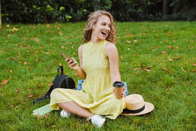 Een lachende positieve vrouw zit buiten op het gras in het natuurpark en houdt koffie vast met behulp van een mobiele telefoon die naar muziek luistert met een koptelefoon.