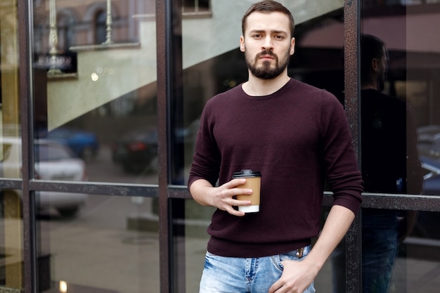 Een lachende trendy man met een hoed, wandelen in de stad een kopje koffie in de hand. Afbeelding van een lachende man van 30 met een bril die door de straat loopt en afhaalkoffie vasthoudt