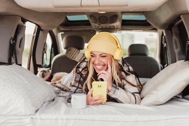 Een lachende vrouw die binnen een minicamper neerlegt met een mobiele telefoon en naar de muziek luistert met een gele koptelefoon Travel vanlife digital nomad people