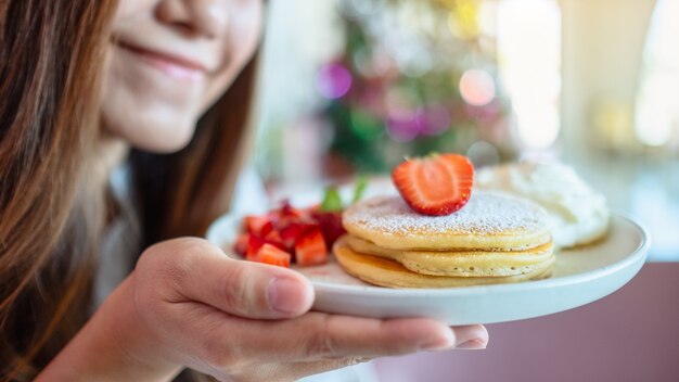 Een lachende vrouw die een bord pannenkoeken met aardbeien en slagroom vasthoudt en toont