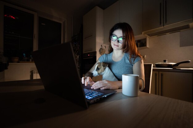 Een lachende vrouw met een bril zit achter een draadloze computer in de keuken met een puppy van Jack Russell Terrier op haar knieën Studente 's nachts studerend voor het examen en koffie drinkend