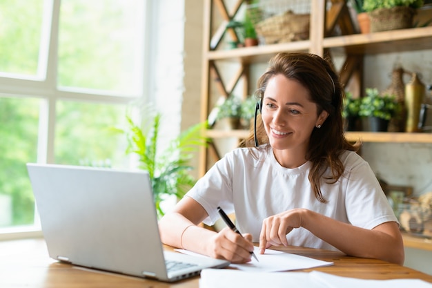 Een lachende vrouw werkt thuis met papieren voor een laptopmonitor, ze controleert papieren tijdens...