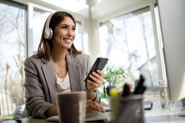 Een lachende zakenvrouw met koptelefoon die smartphone gebruikt terwijl ze op de computer in haar thuiskantoor werkt.