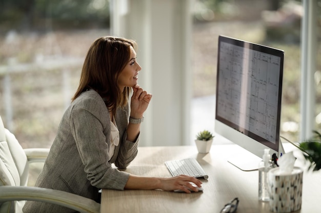Een lachende zakenvrouw zit alleen in haar kantoor aan huis en werkt op de computer.