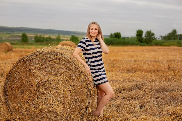 een langharig blond meisje in een lichte zomerjurk leunt tegen een hooiberg in een tarweveld