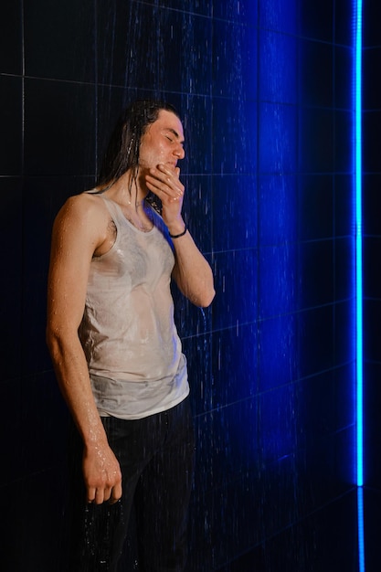 Een langharige man in een nat t-shirt staat in een douche die wordt verlicht door neonlicht