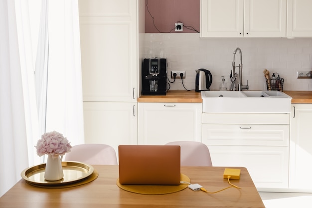 Een laptop en een pot met bloemen op de houten eettafel in de keuken op een zonnige dag