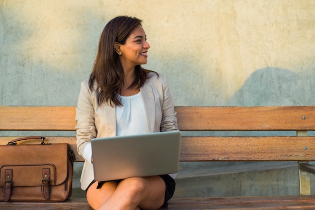 Een latina-vrouw zittend op een bankje met haar computer kijkt glimlachend naar de kopieerruimte