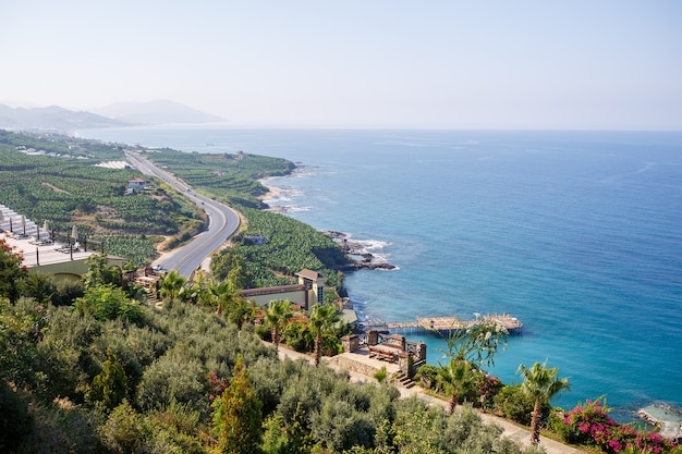 Een lege asfaltweg slingert langs de adembenemende schilderachtige kustlijn op een zonnige zomerdag. Een spectaculaire opname van een kustweg met uitzicht op een heldere blauwe lucht en de kalme Middellandse Zee.