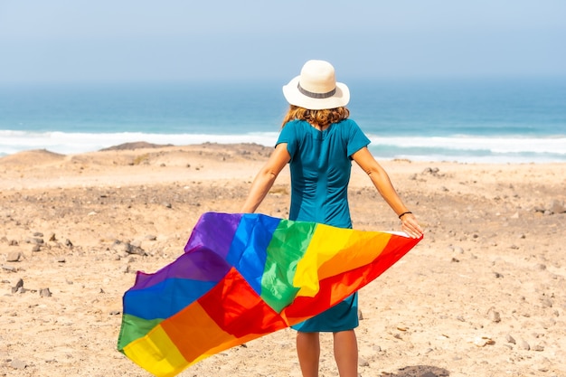 Een lesbisch persoon met een groene jurk, een witte hoed en met de lgbt-vlag bij de zee, een symbool van homoseksualiteit