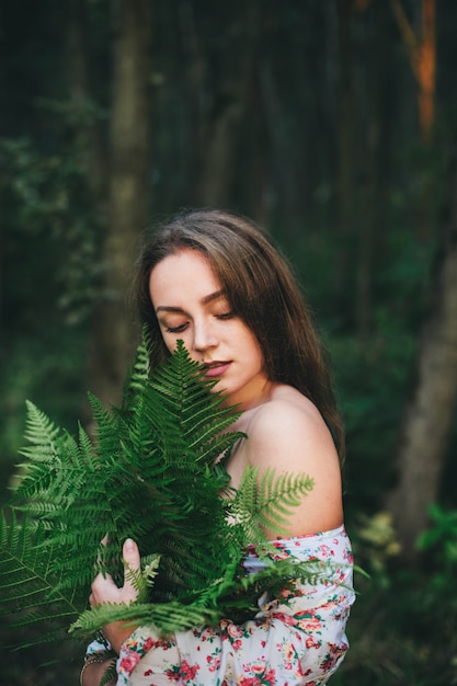 Een leuke vrouw in een bloemenjurk zit met een varenboeket in het bos.