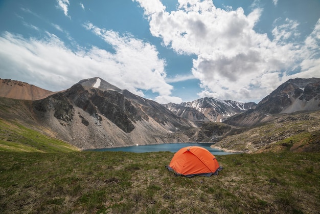 Een levendige oranje tent op een grasrijke heuvel met uitzicht op een bergmeer van schijnblauwe kleur tussen hoge bergen in veranderlijk weer Tent bij een diepblauw meer tussen zonverlichte grote bergen onder bewolkte lucht