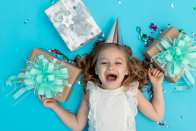 Een lief gelukkig klein meisje in een katoenen witte jurk op een blauwe achtergrond in een feestelijke hoed met geschenken en confetti schreeuwt hoera en lacht van vreugde om haar verjaardag te vieren een plek voor tekst