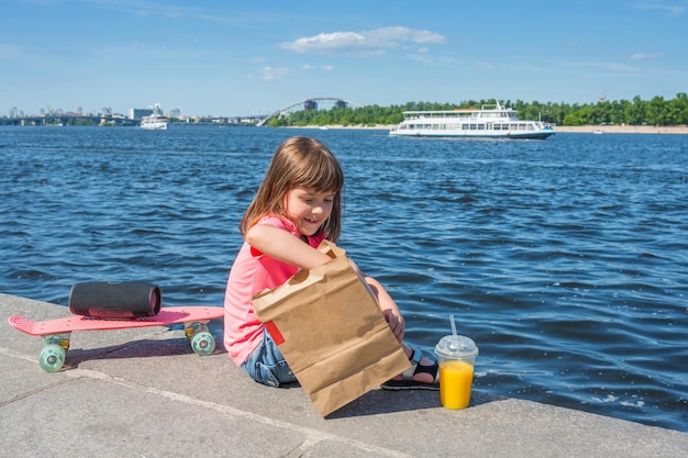 Een lief klein meisje op de achtergrond van een stadsrivier met een zak producten op een zonnige dag