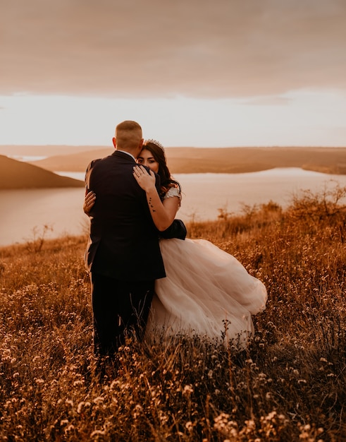 Een liefdevol paar bruiloft jonggehuwden in witte jurk en pak lopen knuffel kussen werveling op hoog gras in zomer veld op berg boven de rivier