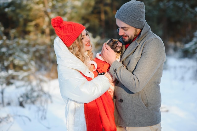 Een liefdevol stel loopt bij de handen op de achtergrond van een besneeuwd winterbos Een man houdt een hond vast Een meisje in een wit donsjack en een rode hoed