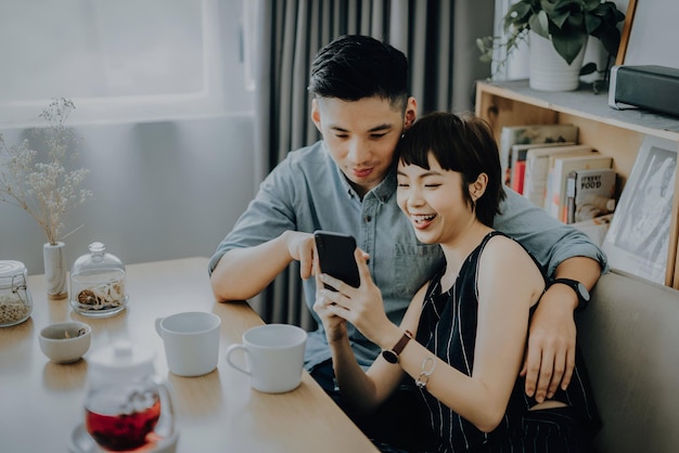 Een liefhebbend jong echtpaar zit op de bank in de woonkamer koffie te drinken en samen naar een smartphone te kijken.