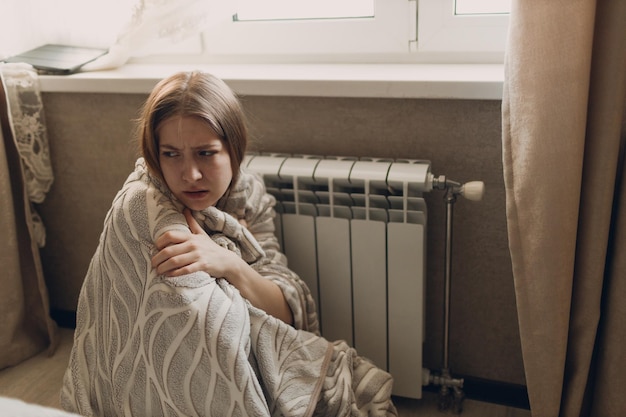 Een lijdende vrouw in een plaid zit bij de verwarmingsbatterij in het huis en bevriest