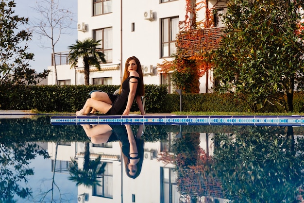 Een luxe aantrekkelijke vrouw in een zwarte jurk rust bij het blauwe zwembad, geniet van een rijk leven