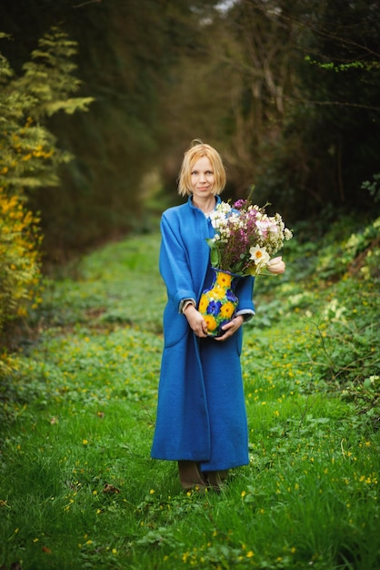 Een magere vrouw van volledige lengte in een blauwe jas staat in een groen bos. Ze houdt een vaas vast met een boeket verschillende bloemen Verticaal frame