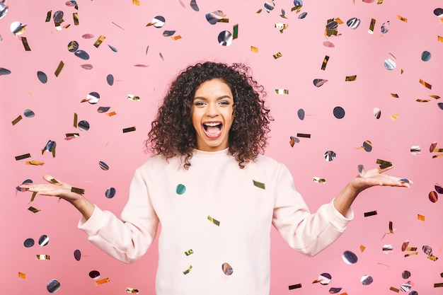 Een magische tijd - portret van een heel gelukkig Afrikaans Amerikaans jong meisje met armen uit, lachend naar confetti vallen geïsoleerd tegen roze.