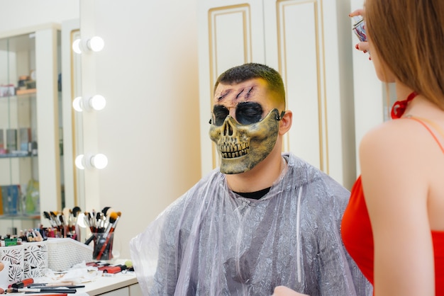 Een make-up specialist meisje maakt een feestelijke griezelige make-up voor een man voor de Halloween-vakantie in de studio. Gelukkig Halloween-concept.