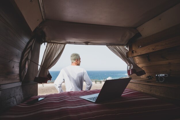 Een man alleen geïsoleerd van de samenleving met zijn minivan-camper die reist en geniet van vakanties - digitale nomade mensen levensstijl werken met laptop kijkend naar de zee - reis en vakantie buitenshuis