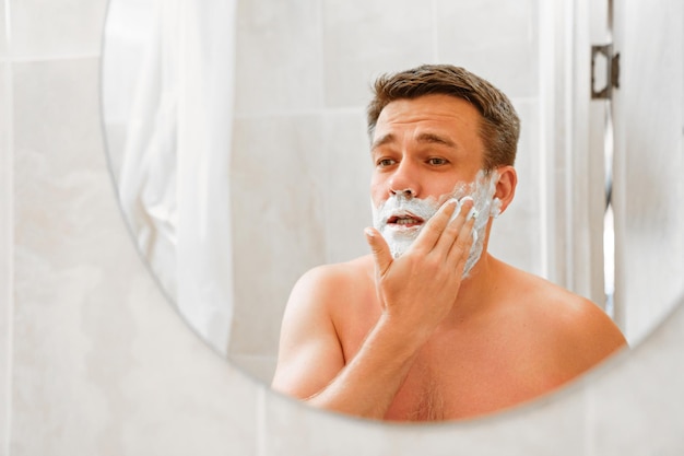 Foto een man brengt scheerschuim aan op zijn gezicht en kijkt in een ronde spiegel