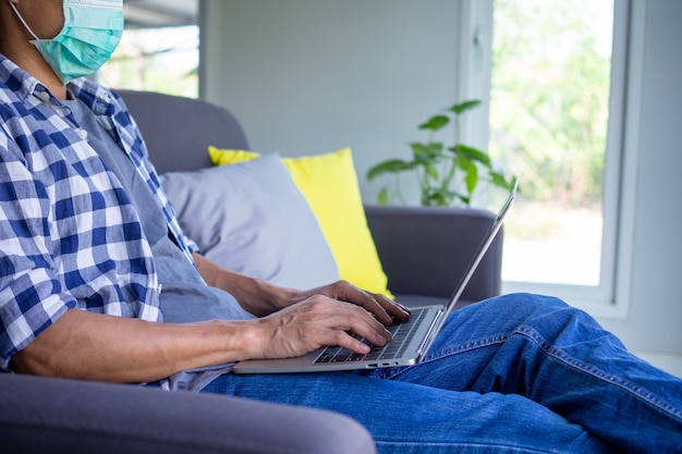 Een man die een masker draagt, gebruikt thuis een laptop om online te werken of te studeren om de verspreiding van het virus te voorkomen. Thuiswerken, online concepten bestuderen