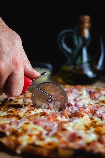 Een man die een pizza snijdt met een pizzasnijder. er zijn oregano en olijfolie op de achtergrond. (verticaal)