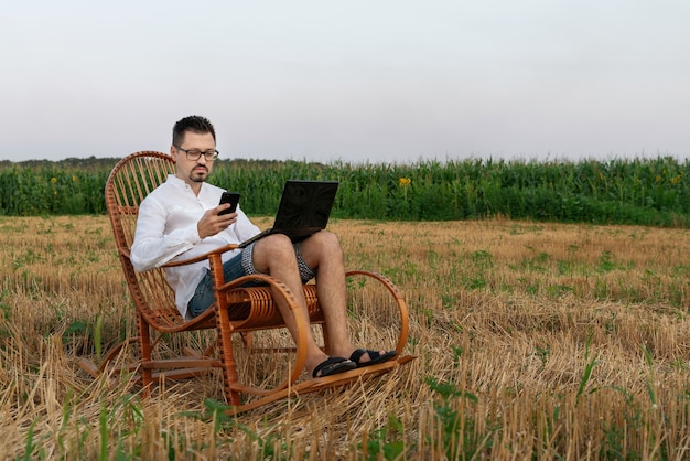 Een man die een smartphone vasthoudt terwijl hij op een laptop werkt en in een schommelstoel midden in een veld zit