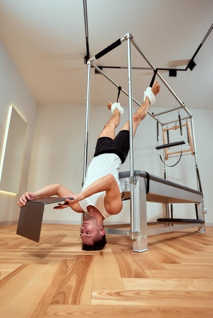 Een man die pilates doet, zich strekt en aan een computer werkt op één been op een reformerbed in de sportschool