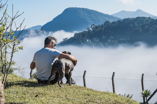 Een man en een Pit Bull-hond bewonderen de natuur en de grote mist in de bergen van PetrÃ³polis, Brazilië, die de stad onder de wolken bedekt. Liefdevolle relatie tussen mens en dier.