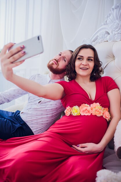 Een man en een zwangere vrouw maken selfie zittend op de bank. ze houden van elkaar. ze zijn thuis.