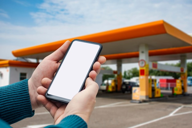 Een man houdt een close-up van een smartphone met een wit scherm in zijn handen tegen de achtergrond op een tankstation Technologiemodel voor apps en websites