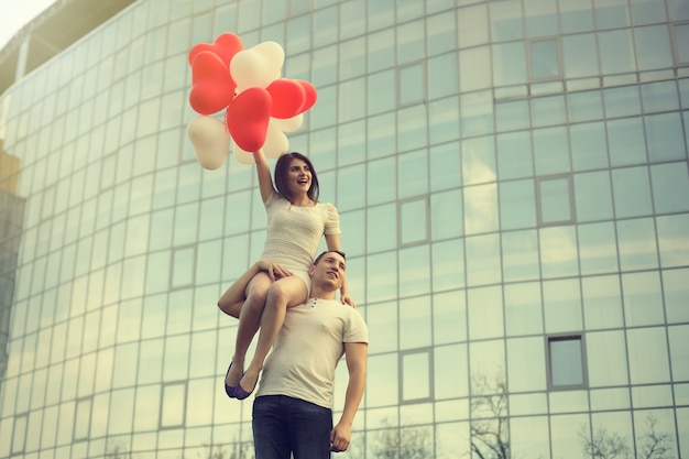 Een man houdt een meisje met ballonnen op zijn schouder tegen de achtergrond van een modern gebouw.