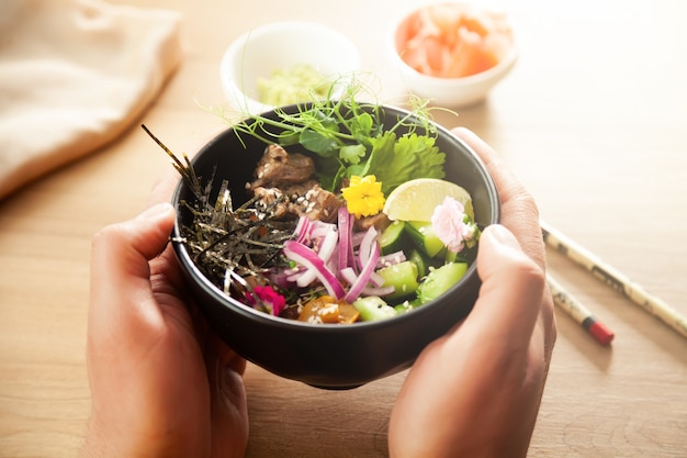 Een man houdt een Poke-salade met rundvlees in een kom in zijn handen. Ingrediënten Rundvlees, Nameko champignons, cherrytomaatjes, rijst, komkommer, rode ui, sesamzaad, koriander, limoen. Aziatisch saladeconcept.
