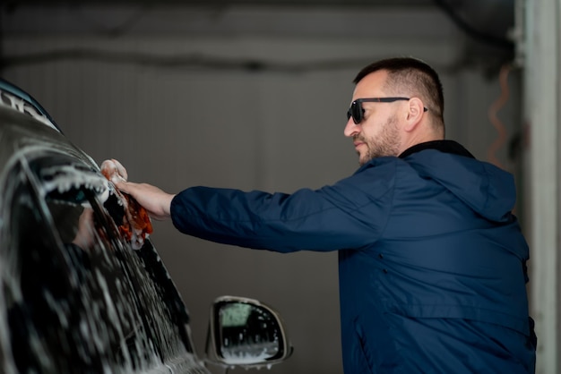 Een man in een blauwe jas en zonnebril wast zijn auto bij een selfservice carwash