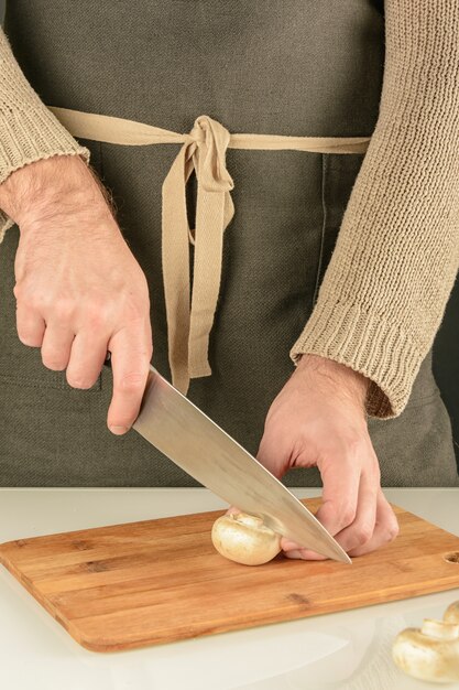 Een man in een donkere schort snijdt een champignon op een houten bord.