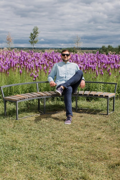 Een man in een hemd en broek zit op een bankje in een park bij een bloemenveld