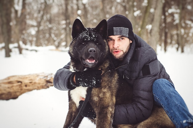 Een man in een jas en een gebreide muts loopt door een besneeuwd bos met een Amerikaanse Akita-hond
