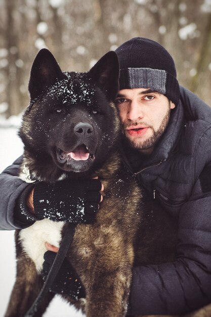 Een man in een jas en een gebreide muts loopt door een besneeuwd bos met een Amerikaanse Akita-hond